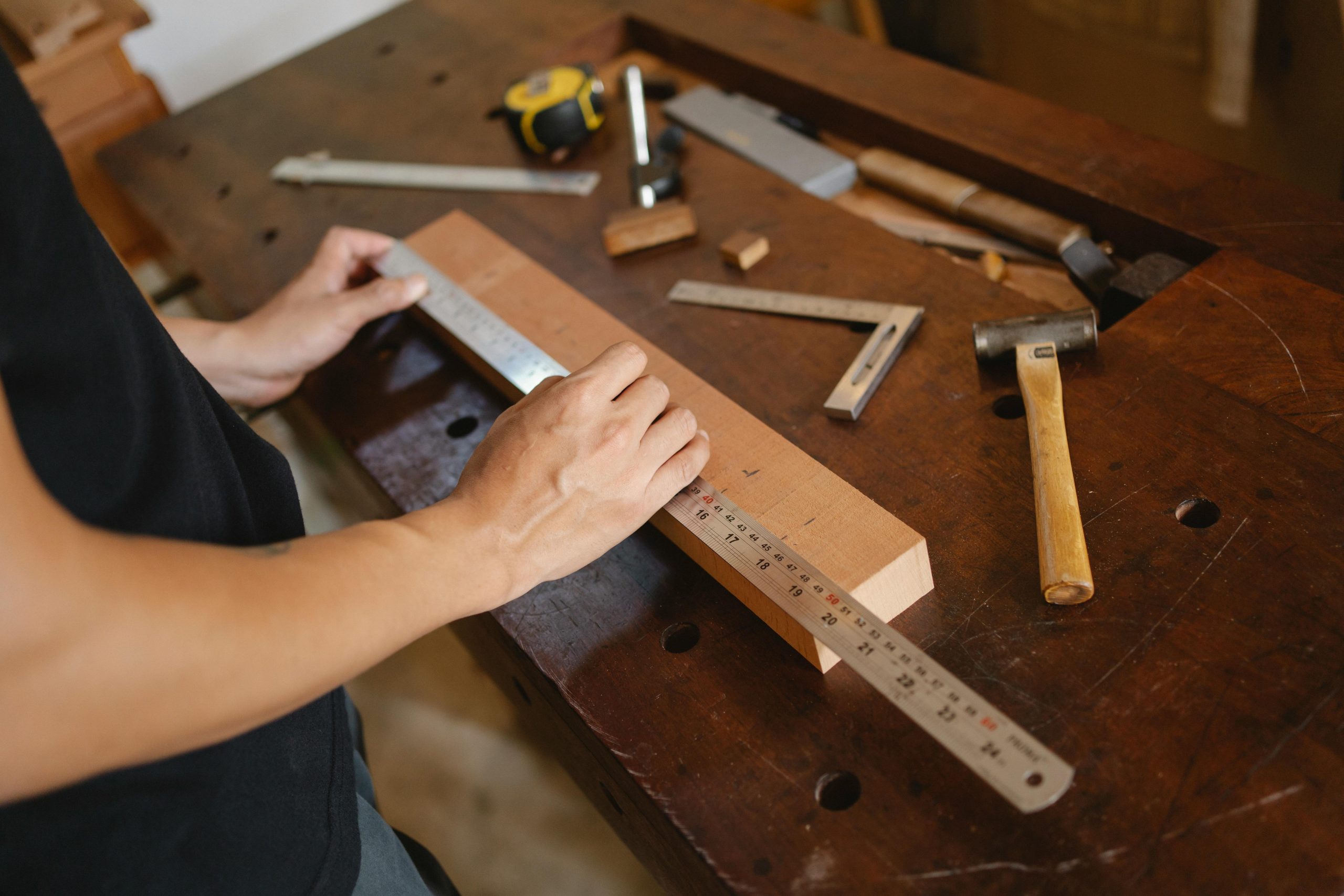 From above of crop anonymous male measuring timber plank attentively on table with hammer in workshop